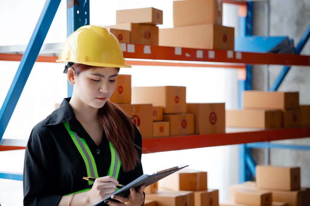 Asian female worker wearing safety clothing is in a warehouse, she is checking the goods in the ware